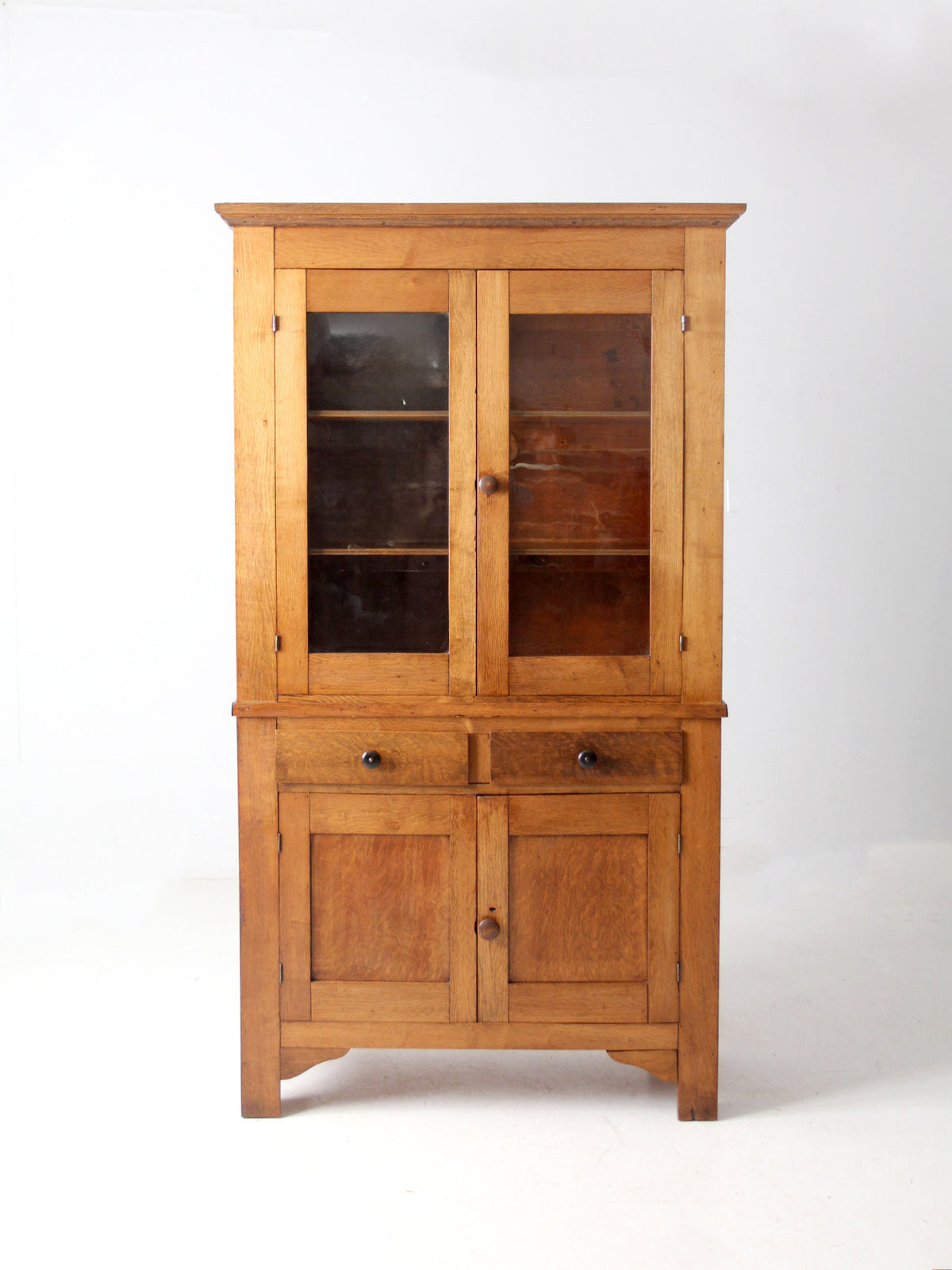 An antique wooden hutch cabinet with a warm honey-toned finish, featuring two upper glass-front doors, two lower paneled doors, and two center drawers with dark metal knobs, photographed against a plain white studio background.