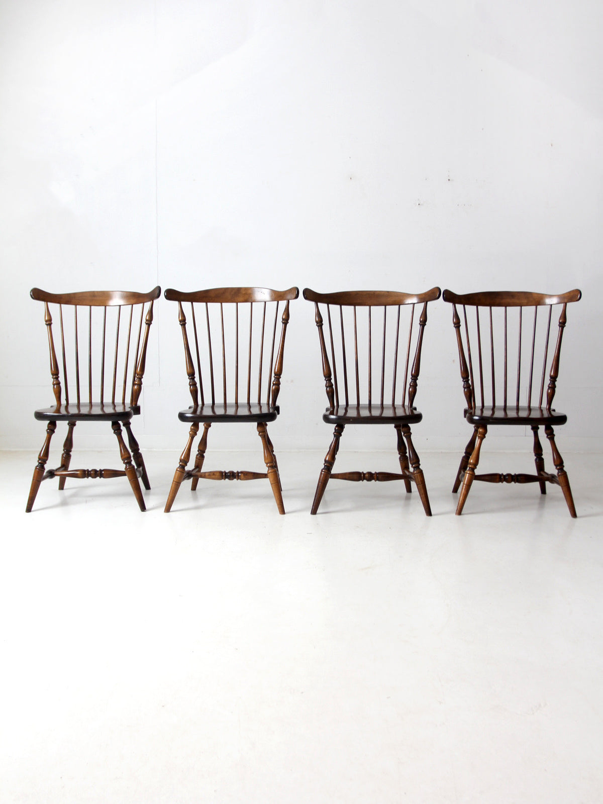 Four dark brown wooden spindle-back chairs arranged in a row against a plain white studio background.