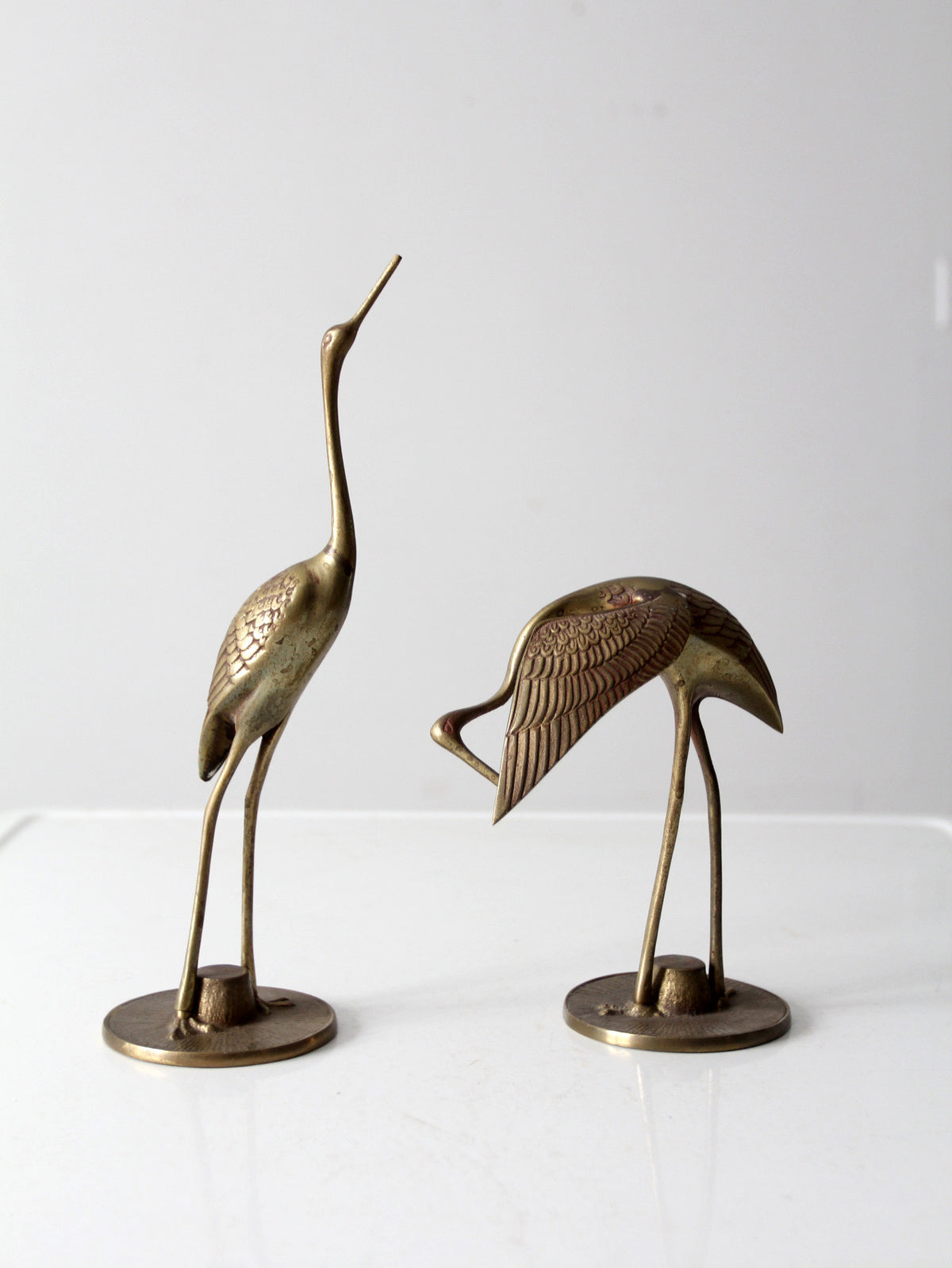 Two brass cranes on round bases, one standing tall and the other bent forward, photographed against a plain light background.