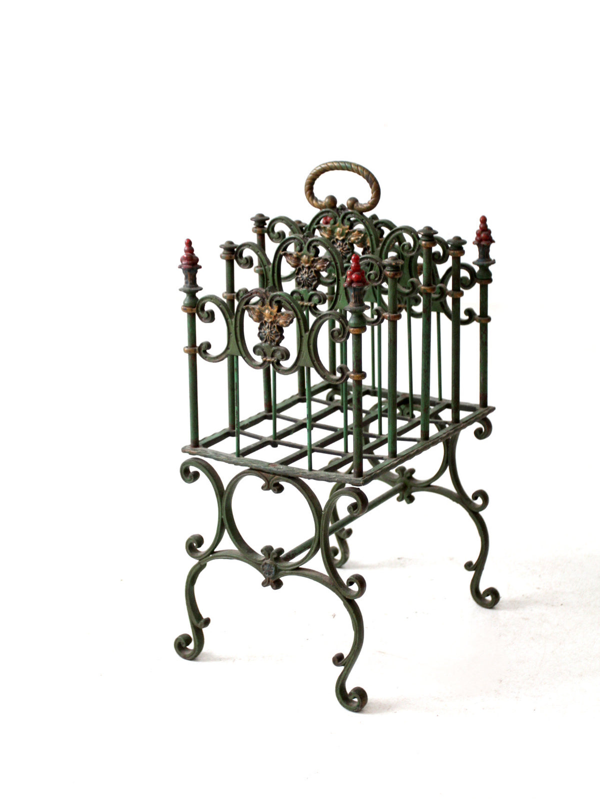 An ornate antique cast iron magazine basket with a dark green patina, red painted finials, and gold-toned decorative accents, shown on a plain white background.