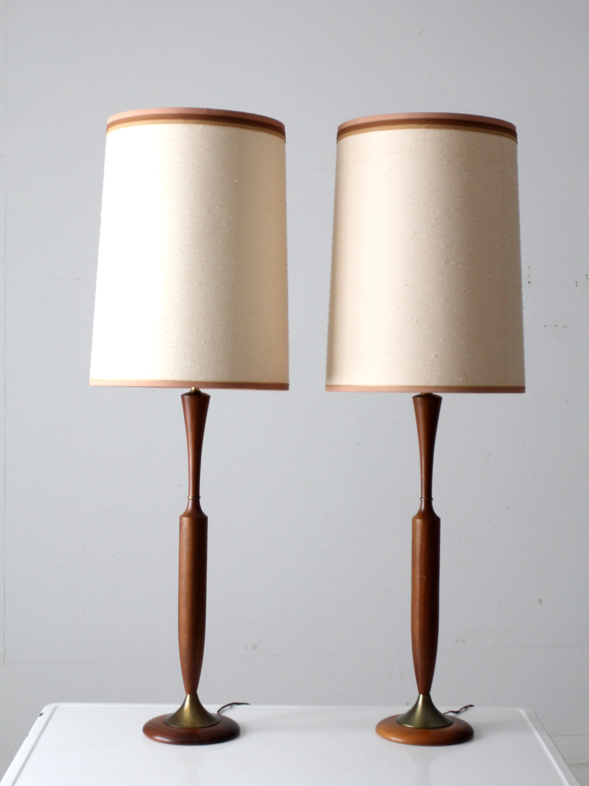 A pair of mid-century modern table lamps with tapered beige fabric shades and sculptural dark brown wooden bases, displayed on a white surface against a light gray wall.
