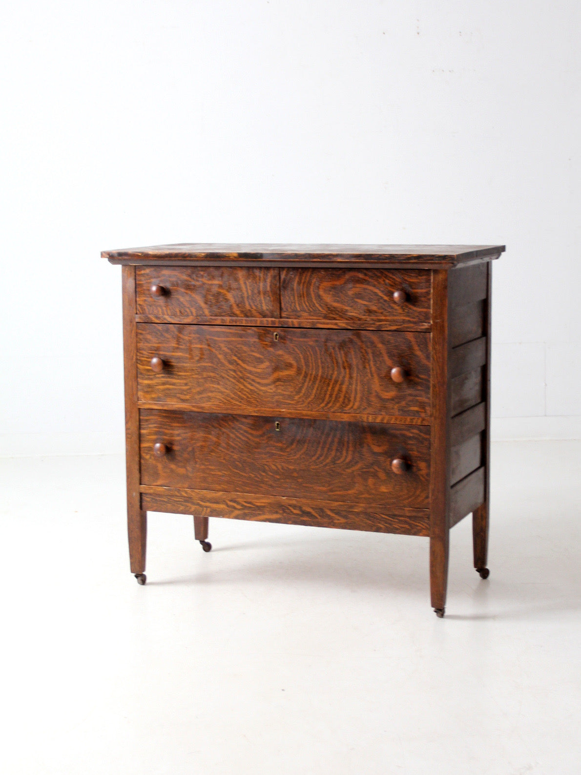 An antique three-drawer wooden chest of drawers with a rich dark brown grain pattern, small round knobs, and casters, photographed against a plain white studio background.