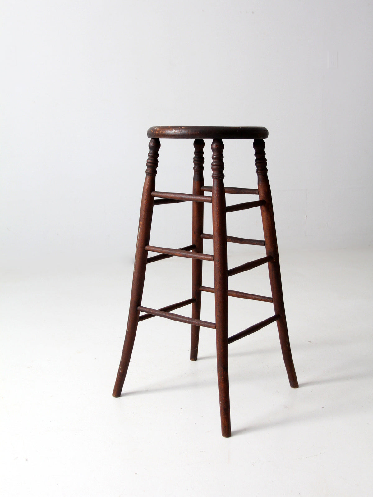 An antique dark brown wooden stool with a round seat, turned spindle legs, and front support rails, photographed against a plain white studio background.