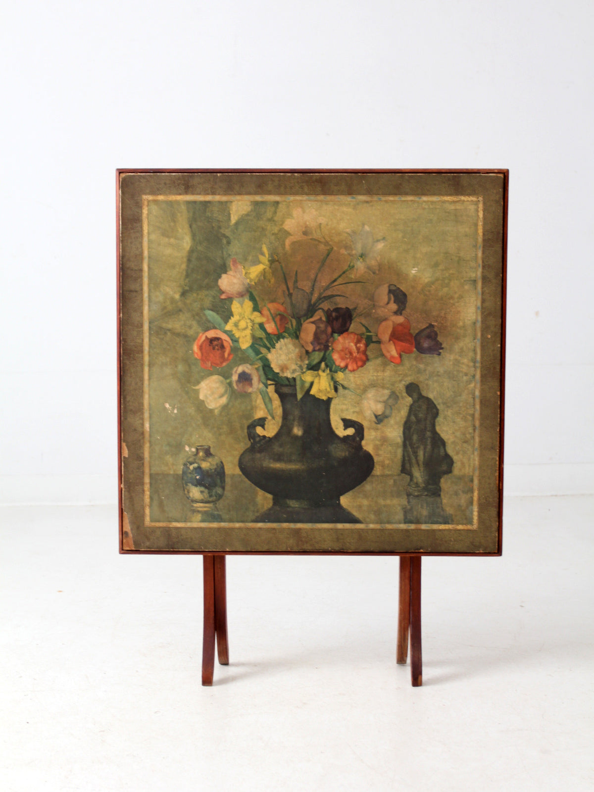 A vintage tilt-top table with still life painting depicting a bouquet of red, yellow, and white flowers in a dark vase against a muted olive background.