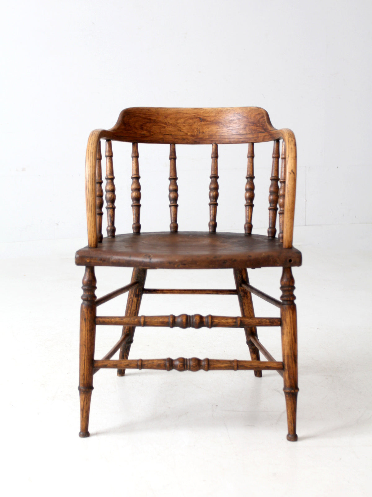An antique wooden spindleback captains chair with a curved backrest, turned spindles, and a round seat, shown against a plain white studio background.
