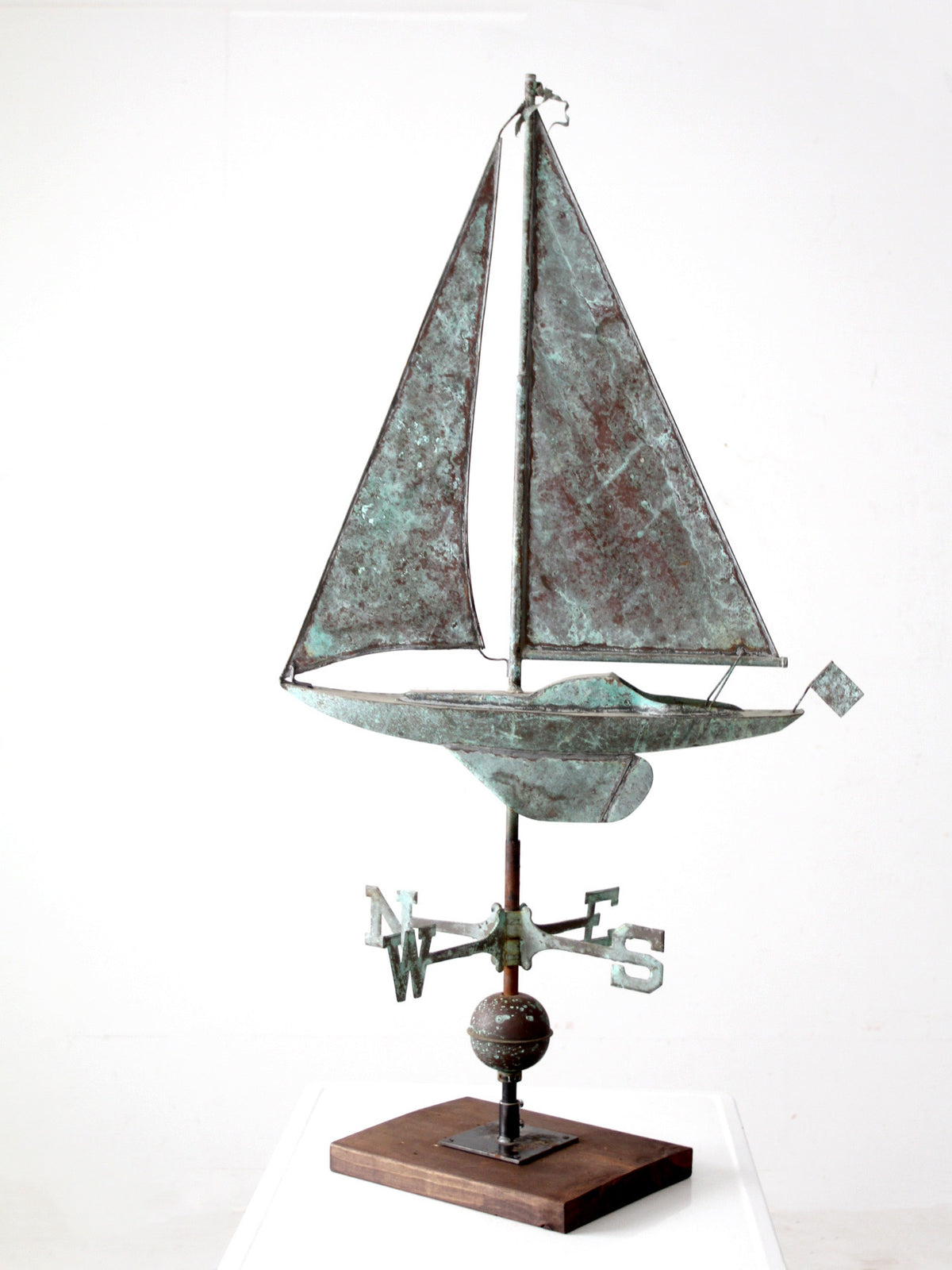 A vintage decorative metal sailboat  weathervane with a weathered greenish-bronze patina, mounted on a wooden base with a compass and finial detail, photographed against a plain white studio background.