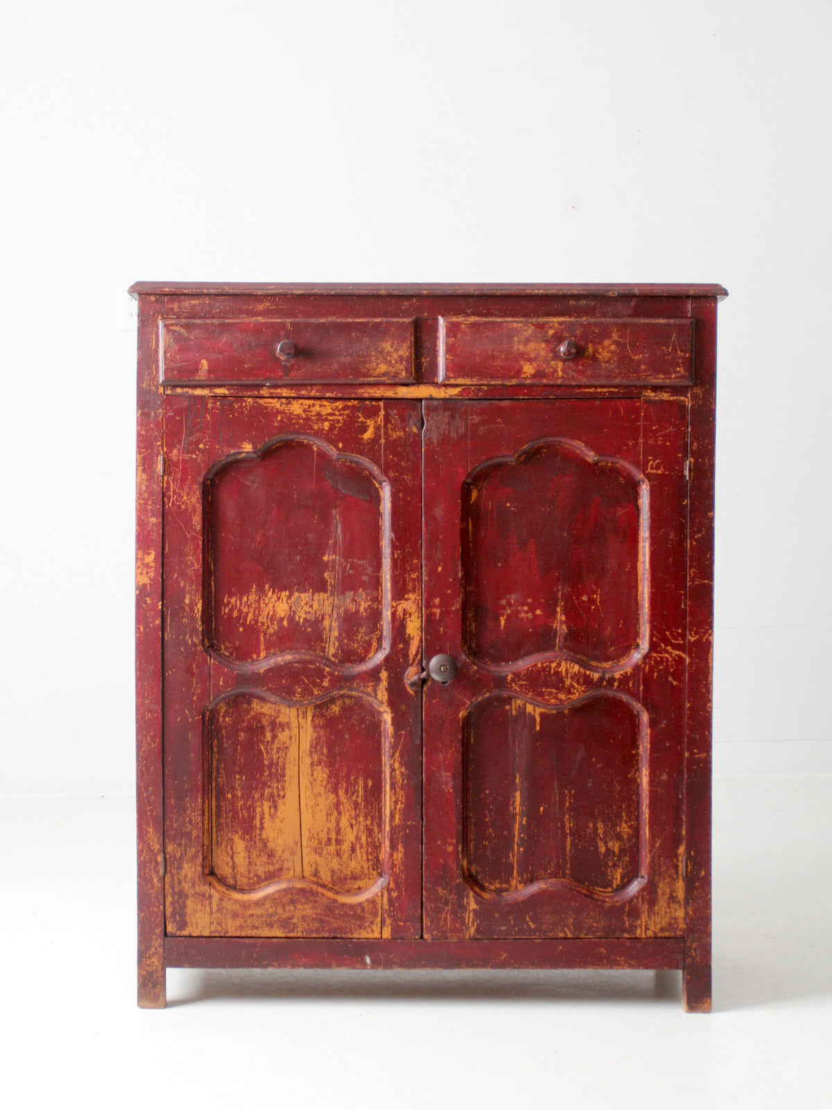 An antique  wooden cabinet with distressed finish revealing a warm brown wood underneath, featuring two doors with decorative arched panels and two small drawers at the top.