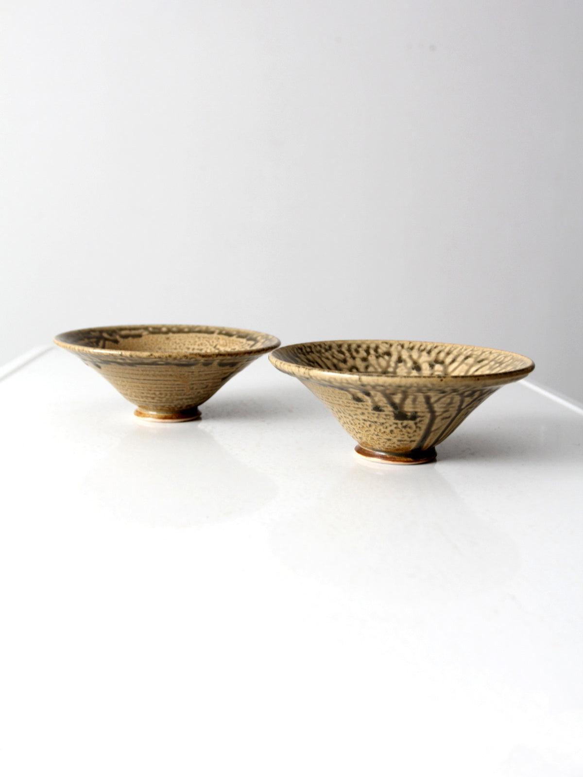 Two handmade ceramic bowls with a textured, speckled beige and brown glaze, displayed on a reflective white surface against a light gray background.