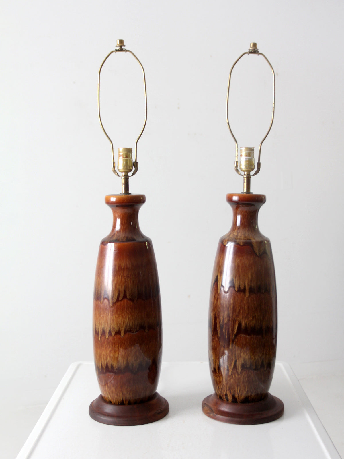 A pair of vintage ceramic table lamps with glossy brown and amber glazed bodies, brass metal shades, and dark wooden bases, set against a plain white background.