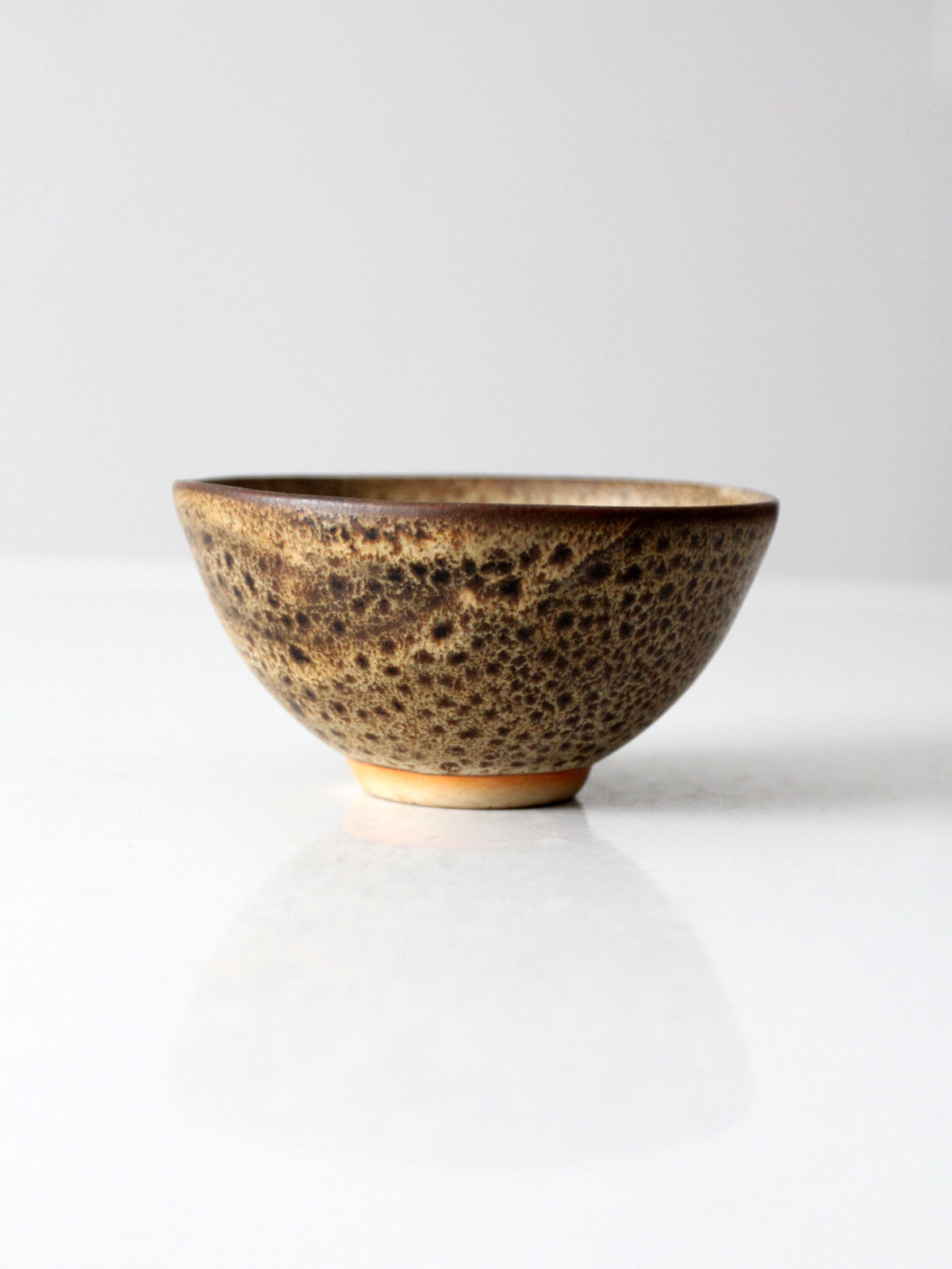 A handmade ceramic bowl with a textured, speckled brown and beige glaze, standing on a reflective white surface against a light gray background.