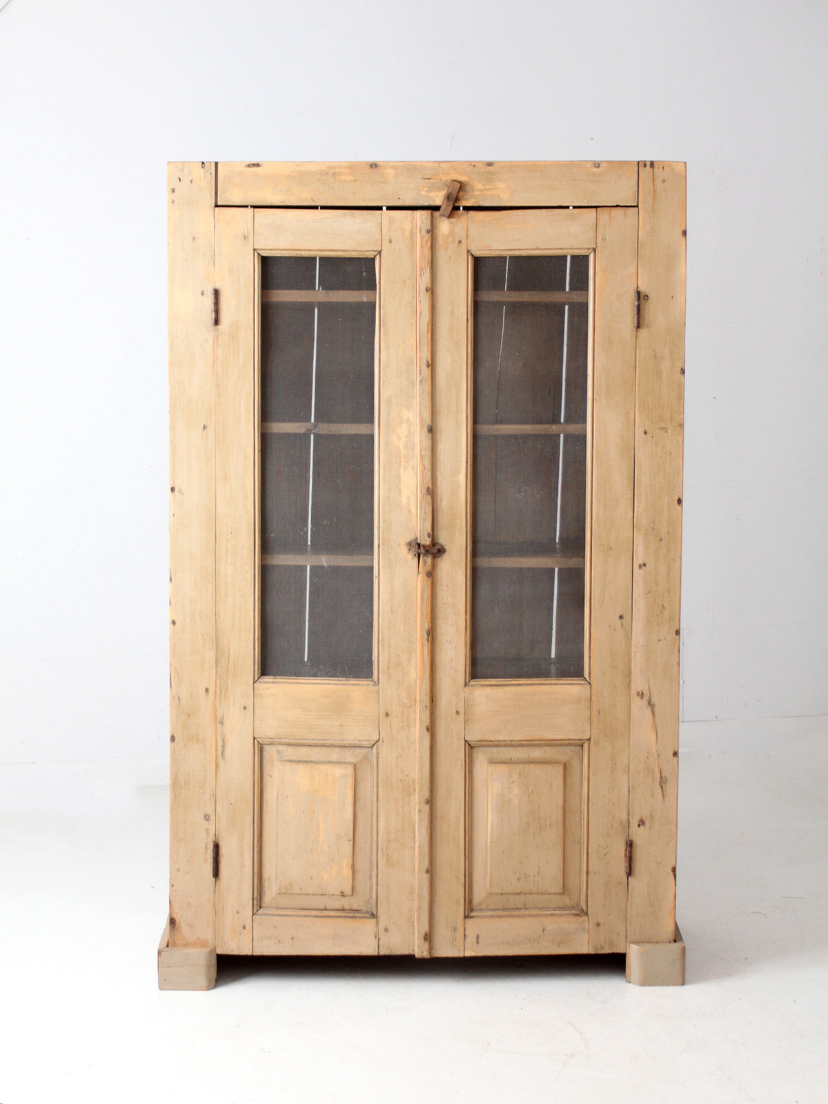 An antique rustic light wood cabinet with double doors featuring screen panes and dark metal hinges, photographed against a plain white studio background.