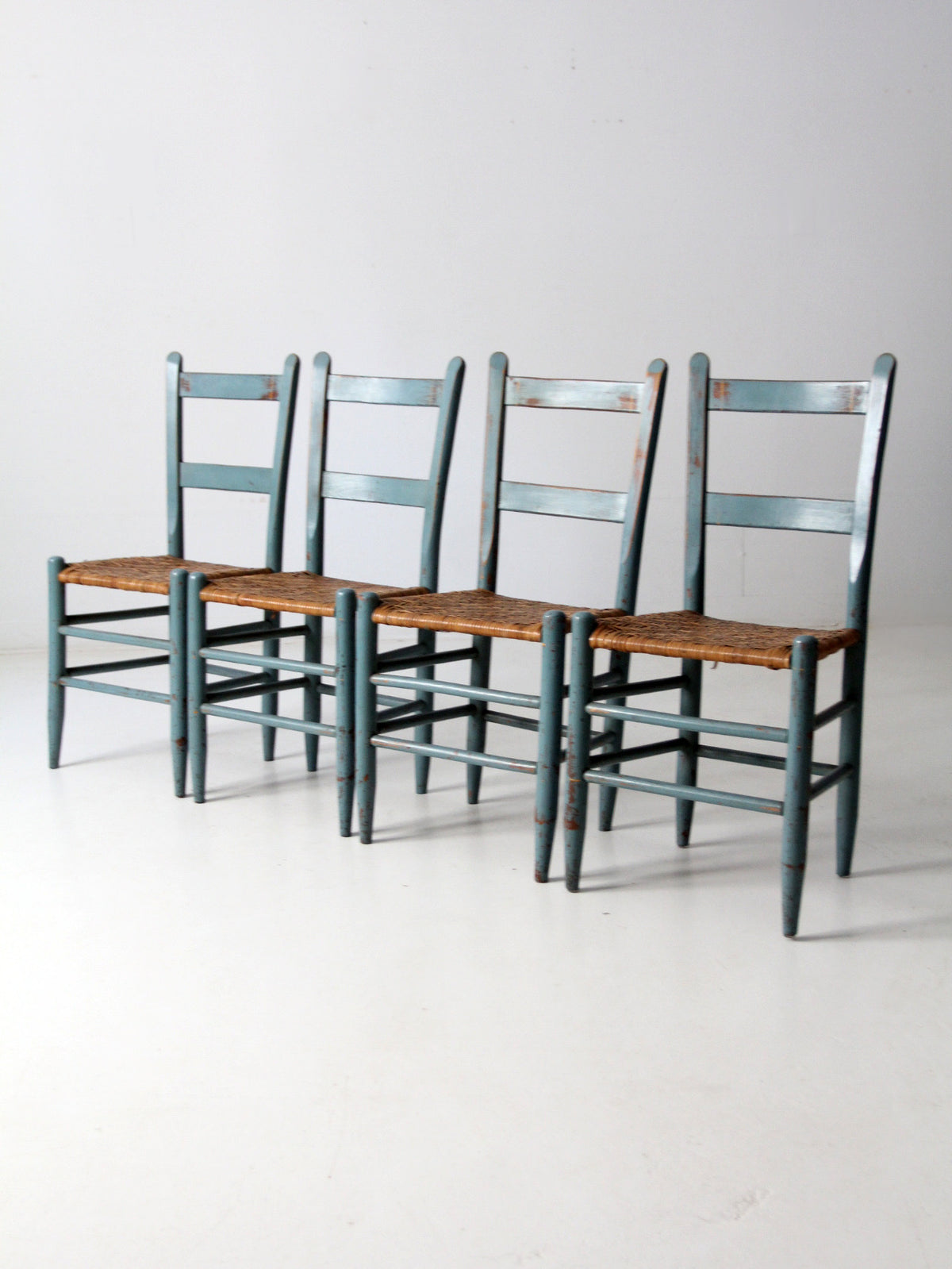 Four matching antique wooden dining chairs with distressed teal-blue painted frames and natural brown woven seats, arranged in a row against a plain light gray studio background.